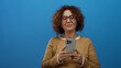 © Krakenimages.com - Middle aged woman with curly hair in glasses using smartphone against a blue background wall, wearing brown sweater, showing thoughtful expression and hispanic ethnicity.