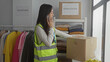 © Krakenimages.com - Woman volunteering indoors at a charity center, wearing a reflective vest, talking on the phone beside donation boxes and clothing racks.