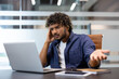 © Tetiana - Close-up photo of a young Indian man sitting at an office desk, looking angrily at the laptop screen and having an emotional conversation online