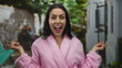 © Krakenimages.com - Smiling young hispanic woman in a pink robe outside pointing both ways, surrounded by lush greenery and a relaxed outdoor setting, exuding happiness and youthful energy.
