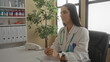 © Krakenimages.com - Hispanic woman doctor in clinic wearing stethoscope seated at desk in medical workplace with plants and documents on table suggesting professional healthcare setting.