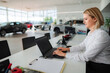 © Jelena - Car saleswoman typing on laptop in automotive dealership