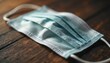 © Vadym - Light blue surgical face mask rests on textured wooden planks. Close view shows mask folds and ear loops. Object suggests health safety and protection measures in daily life.
