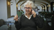 © Krakenimages.com - Elderly woman holds phone to ear and makes a fist gesture inside a building cafe with arched seating and lamps; joy connection.