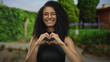 © Krakenimages.com - Woman smiling and forming heart shape with hands in a lush green park outdoors.