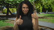 © Krakenimages.com - Woman with curly hair poses in a sunny park, expressing confidence and wearing glasses, showing two thumbs up amid a lush green outdoor setting, embodying young hispanic latina energy.