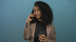 © Krakenimages.com - Woman with curly hair in glasses holding coffee cup and talking on smartphone against isolated blue wall background, conveying business communication.