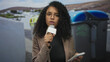 © Krakenimages.com - Woman reporter holding microphone in front of recycling containers on outdoor street setting, conveying environmental concern with intense expression.