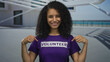 © Krakenimages.com - Woman smiling in outdoor setting wearing volunteer shirt, pointing to text while standing with building background and ocean view, showcasing community involvement.