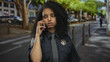 © Krakenimages.com - Woman police officer in uniform talking on phone standing on street, outdoors setting with trees and blurred background suggesting an urban environment.