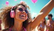 © miss irine - Young woman enjoys music on headphones at beach party. She wears sunglasses and bikini smiling with raised arm. People relax on sunny day at seaside with balloons. Friends have fun in background.