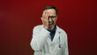 © Krakenimages.com - Mature doctor smiling and covering one eye with hand against a vibrant red background.