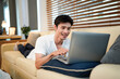 © Stella 16 - Confident Asian man using laptop while lay down on cozy sofa at modern home