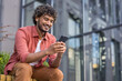 © Tetiana - Smiling young Indian man sitting on a bench outside wearing headphones and using his mobile phone