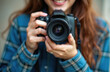 © Viktor - Young woman smiles holding DSLR camera ready to shoot. Girl wears blue checkered shirt, focusing lens. Capturing moments with hobby and passion for photography.