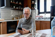 © lordn - Smiling senior man sitting at a kitchen table, using his smartphone with a glass of water beside him in a cozy home setting