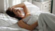 © wedmoments.stock - Young woman with brown hair, smiling while waking up in a cozy bedroom, surrounded by soft pillows and blankets, enjoying a peaceful morning moment of relaxation and comfort