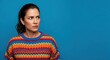 © SimpleDesignStudio - Pensive young adult woman with a furrowed brow looking sideways, dressed in a bright multi-colored knit sweater against a plain blue studio backdrop.