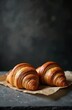 © Maryna - Two golden brown croissants with flaky layers on brown parchment paper on dark stone surface. Fresh baked pastries for breakfast or snack, copy space for product or text.