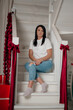 © Татьяна Зернова - A young Caucasian woman with long dark hair sits on a staircase. She wears a white t-shirt and blue jeans. The staircase is decorated with red ribbons and presents.