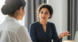© abu - Two women in a modern office are engaged in conversation at a desk, bathed in natural light. One wears a white shirt, while the other sports a navy shirt with white polka dots.