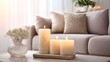© phy - Three lit candles on a wooden tray, with a beige sofa and pillows in the background.