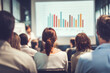 © Photoenthusiast82 - Rear view of a diverse group of business professionals attending a seminar, focused on a presenter explaining charts and graphs on a large projection screen in a bright modern conference room.