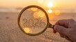 © Vendcat Gemers - A close-up view through a magnifying glass reveals the year 2026 written in the sand, a concept for future planning and new year goals at sunset