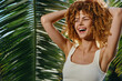 © SHOTPRIME STUDIO - Smiling woman with curly hair enjoys tropical sunlight wearing white tank top under palm leaves, expressing happiness and natural beauty in vibrant outdoor setting.