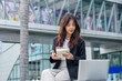 © sorapop - Young Asian woman, busy with business work, wears suit, sits on street, writes in notebook, laptop nearby, embodying professional modern urban lifestyle, dedicated to her tasks in bustling city