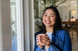 © Wavebreak Media - Woman holding ceramic coffee mug and smiling beside large glass window and potted plant in cafe