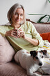 © Tatyana Gladskih - Happy middle aged Woman sitting with Dog on Sofa in living room with cup of tea or coffee