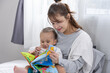 © 220 Selfmade studio - Asian Young mother reading colorful fabric book to baby on bed, tender moment of bonding and early learning through play