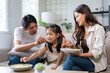 © amnaj - Happy Asian family spending bonding time together, parents feeding their daughter crispy snacks in living room. Cozy domestic scene