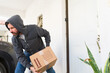 © AntonioDiaz - Criminal man in a hoodie taking a cardboard box from a residential porch, illustrating package theft and home security issues