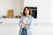 © Prostock-studio - A cheerful young woman with short hair poses in a stylish kitchen, wearing a light blue shirt and jeans. She radiates positivity and comfort in the home setting.