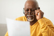 © Prostock-studio - An elderly man wearing a yellow sweater focuses intently as he reads a document. He adjusts his glasses, looking thoughtful and engaged in a comfortable indoor space.