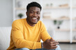 © Prostock-studio - Portrait of successful millennial black guy in casual outfit sitting on couch in cozy living room and smiling at camera, posing alone at home, copy space. Millennials lifestyles concept