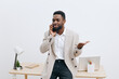 © SHOTPRIME STUDIO - Confident young man with dark skin talking on mobile phone in modern office setting, wearing smart casual outfit, holding hand in gesture, professional workspace background with desk and laptop