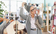 © JackF - Stylish mature woman in glasses is stands in a clothing store and choosing a warm hat against the background of shelf lights. Pensioner buys outerwear