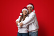 © Prostock-studio - A cheerful couple embraces in front of a bright red background, both wearing warm sweaters and matching Santa hats.