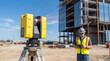© Hiskia - Construction worker using a tablet near a yellow 3D laser scanner on a tripod at a building site. Focuses on modern technology, engineering, surveying, and BIM (Building Information Modeling).