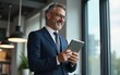 © si - Happy middle aged business man ceo wearing suit standing in office using digital tablet. Smiling mature businessman professional executive manager looking away thinking using modern technology.
