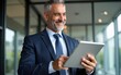 © si - Happy middle aged business man ceo wearing suit standing in office using digital tablet. Smiling mature businessman professional executive manager looking away thinking working on tech device