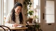 © Таня Андреева - Smiling Asian woman in a cozy sweater writing in a journal at a cafe table by the window with soft natural light.