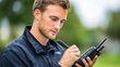 © aMichelle - Young man, wearing a black jacket, is using a two-way radio while taking notes outdoors, surrounded by greenery, demonstrating effective communication in a professional setting