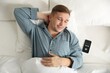 © New Africa - Man lying in bed and smartphone with alarm clock app indoors, top view