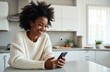 © Vadym - Happy African American woman smiles, using mobile phone in her bright modern kitchen. She holds smartphone, browsing social media, reading online news, or shopping at home.