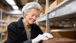 © iuricazac - Woman in warehouse, elderly, smiling, wearing gloves, inspecting boxes on shelf.