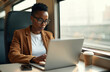© Pete - Young Black woman travels by train, focused on work. Uses laptop computer, smartphone to manage business tasks. Pro stays productive on commute. City environment passes outside window. Coffee cup